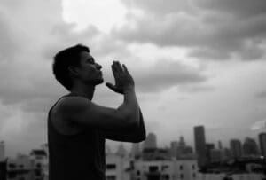 A man standing on a rooftop with hands in a prayer pose, practicing gratitude against a city skyline.