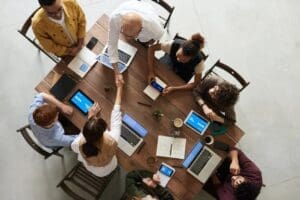 An overhead view of a diverse professional team sitting around a wooden table with laptops and tablets, shaking hands to signify a successful partnership with clients.