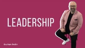 Kristian Livolsi in a pink blazer sitting on a stool against a maroon background, representing modern leadership principles.